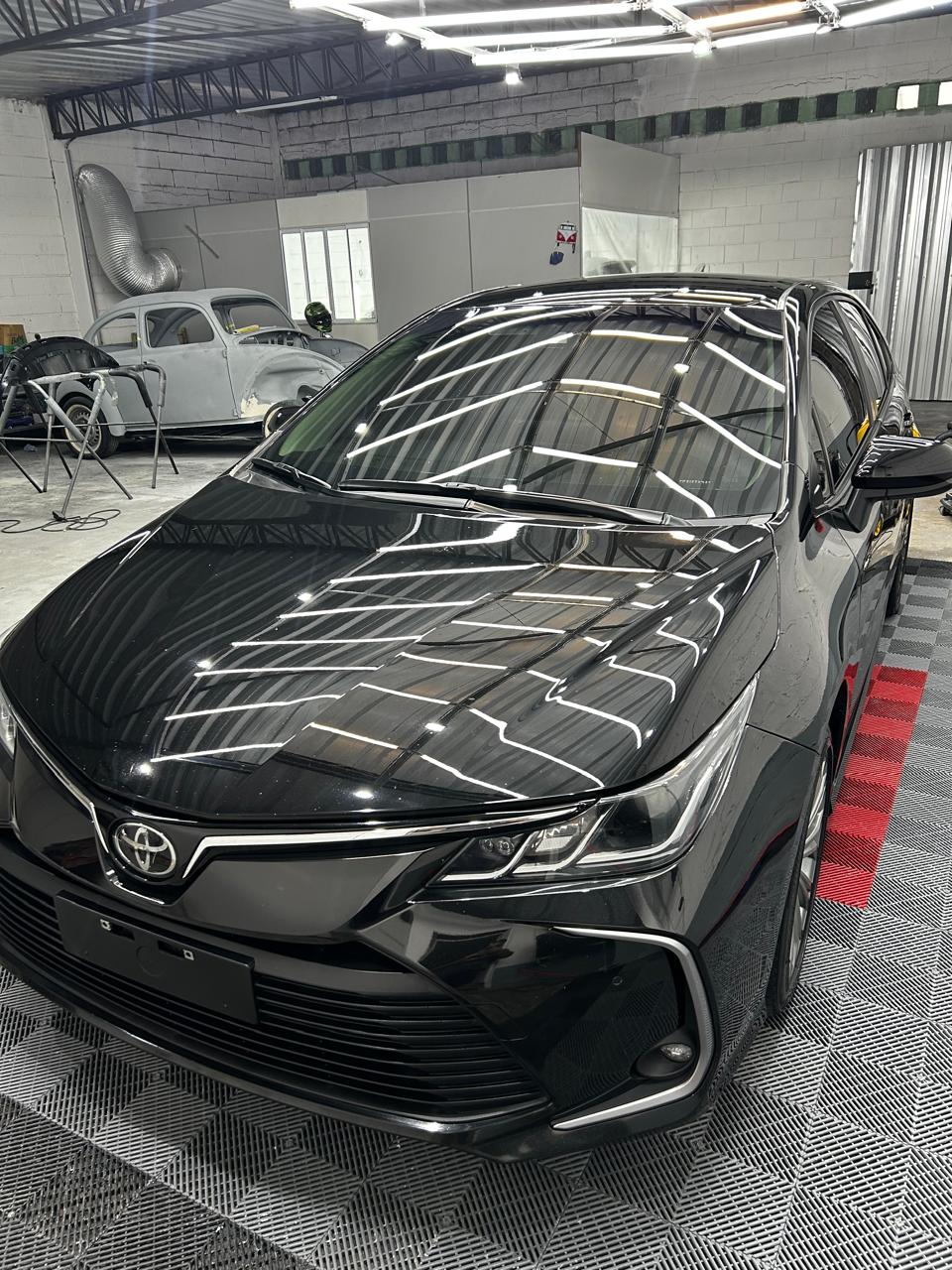 Toyota Corolla preto polido no estúdio, com Fusca em restauração ao fundo